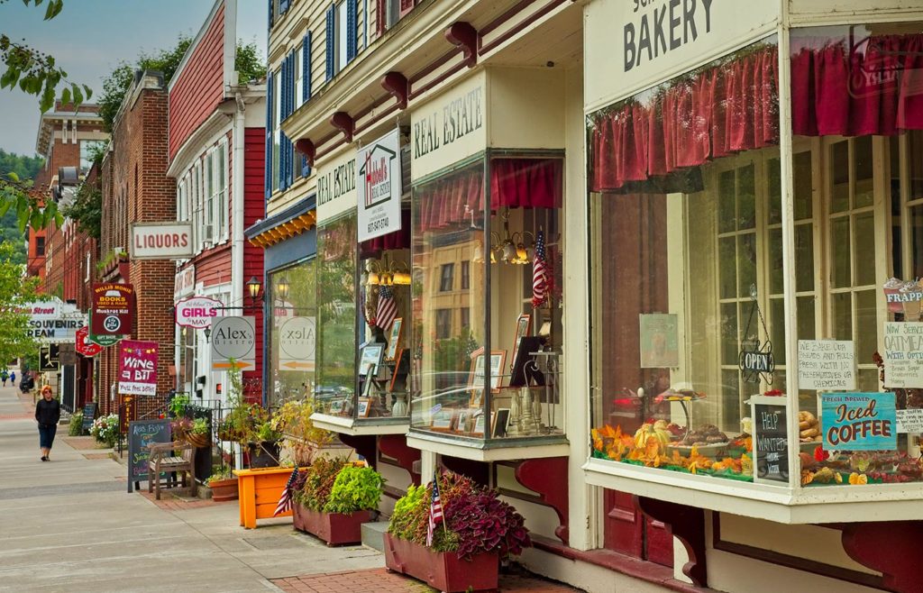 A street-level view of small, connected storefronts with large glass windows along a sidewalk in a town.