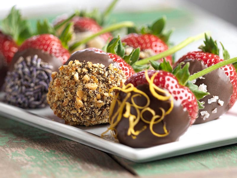 A platter of strawberries dipped in chocolate and topped with various garnishes like nuts, zest, and sea salt.