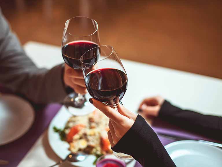 Two people toast with glasses of red wine over a dinner table with plates of food.