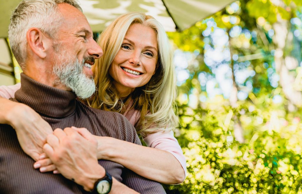 A smiling, older couple with blonde and grey hair pose affectionately, arms wrapped around each other outdoors.