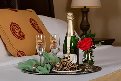 A silver tray on a bed holds champagne, two flutes, chocolate-covered strawberries, and a red rose in a vase.