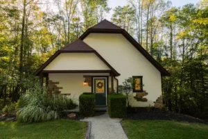 A house with cream walls, brown roof, and stone accents, surrounded by woods, with a front door and a path.