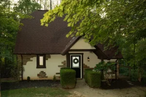 A cozy cottage with a dark shingled roof, cream stucco, and stone accents, nestled among lush green trees.