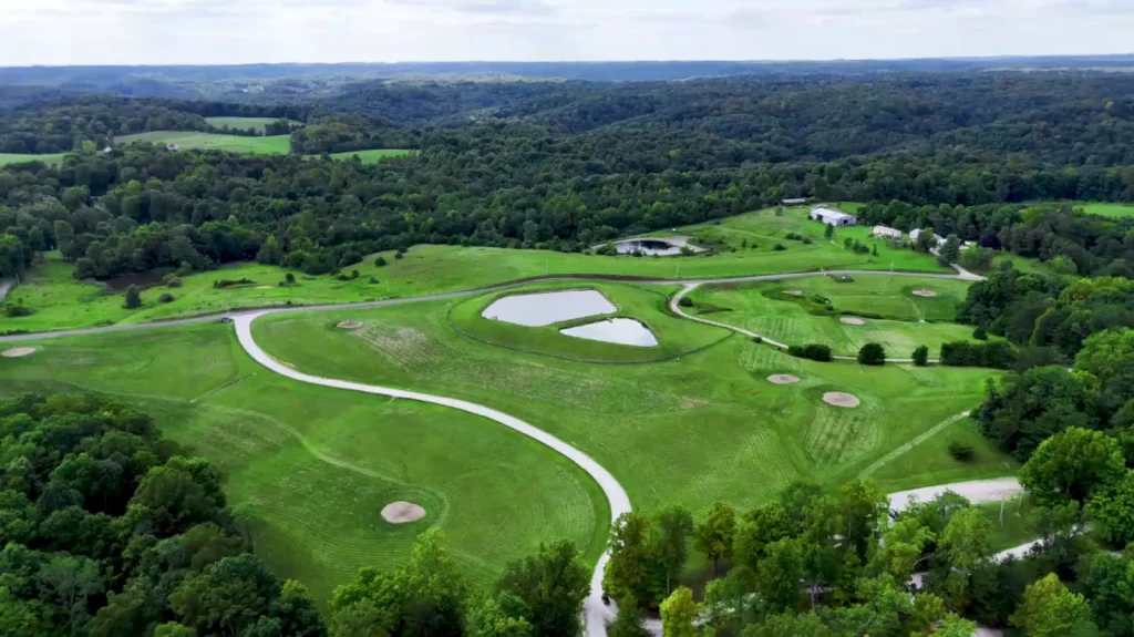 Aerial view of a green, rolling golf course surrounded by dense forest, featuring water hazards and winding paths.