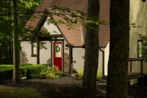 A white cottage with a brown roof, a red front door with a wreath, and stone accents, viewed through trees.