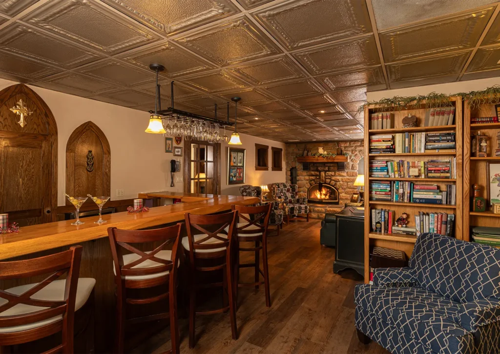 A cozy room with a wooden bar, stools, fireplace, and bookshelves under a patterned tin ceiling.
