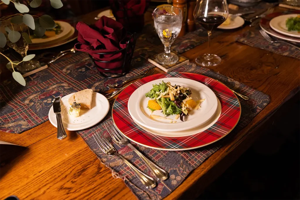 A dinner place setting featuring a salad on a red plaid charger, a small bread plate, and wine glasses on a wood table.