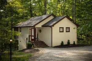A light-colored house with a red door and wooden stairs, nestled in a wooded area with a gravel driveway.