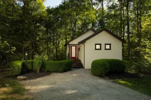 A light-colored small house with a red door stands in a dense forest, featuring trimmed hedges and a gravel driveway.