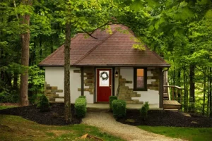 A cottage with a brown roof and stone-accented walls, centered by a red door, sits in a lush, wooded environment.
