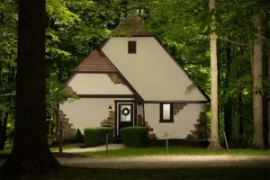 A light-colored, cottage-style house with stone accents and a steep roof, nestled in a dense, green forest.